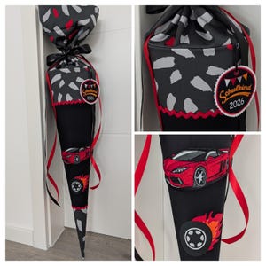 Könnte beinhalten: Eine schwarze und rote Schultüte mit einem Autodesign. Der Kegel hat eine Applikation eines roten Sportwagens, einen Reifen mit Flammen und einen oberen Bereich mit einem grau-weißen Muster. Er hat ein Etikett mit der Aufschrift "Schulkind 2026".