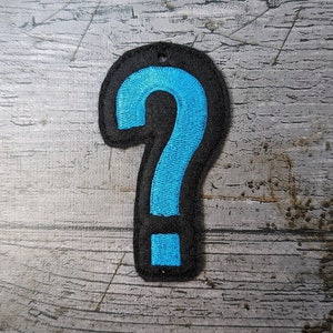 Puede incluir: Un signo de interrogación azul bordado en un parche de fieltro negro.