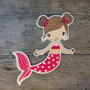 May include: A fabric patch depicting a cartoon mermaid with brown hair in two buns, decorated with pink bows. The mermaid has a pink and white polka-dot tail and a pink top. The patch is outlined in white.