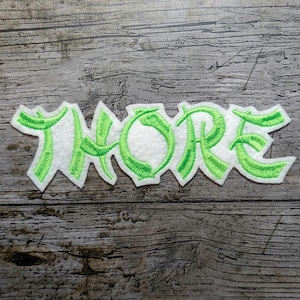 May include: Embroidered patch featuring the word "THORE" in a stylized font. The letters are outlined in bright green stitching and filled with white fabric. The patch has a decorative edge, set against a weathered wood background.