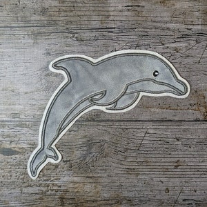 May include: A gray felt dolphin applique with a white outline. The dolphin is leaping out of the water with its mouth open.
