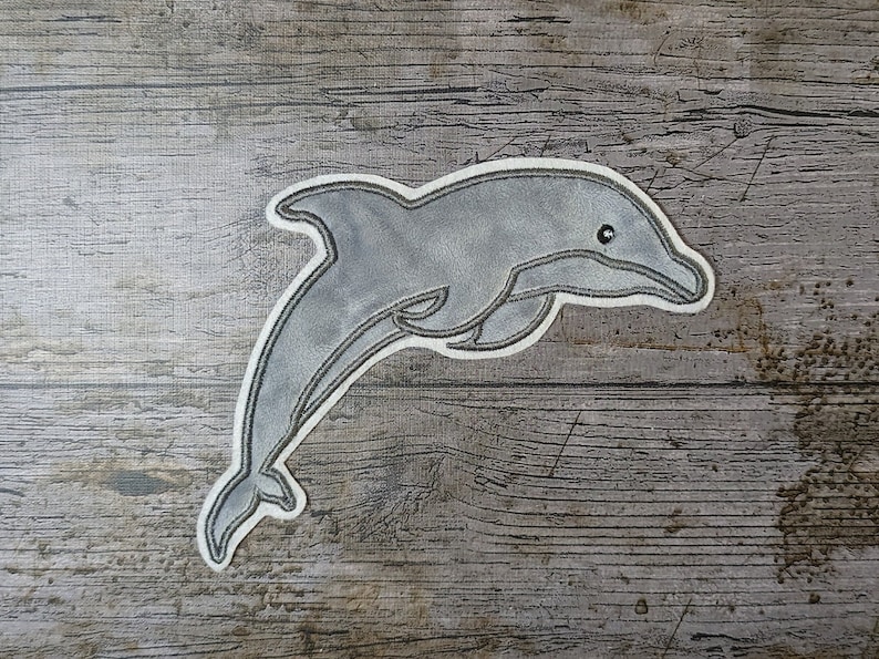 May include: A gray felt dolphin applique with a white outline. The dolphin is leaping out of the water with its mouth open.