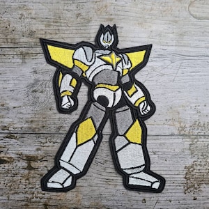 Puede incluir: Parche bordado para planchar con un robot blanco y amarillo con una estrella en el pecho. El robot tiene una cabeza puntiaguda y está de pie con las piernas separadas.