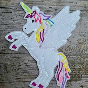 Peut inclure: Une applique en feutre blanc en forme de licorne avec une crinière et une queue arc-en-ciel. La licorne a des ailes et une corne avec un bandeau rose et vert.