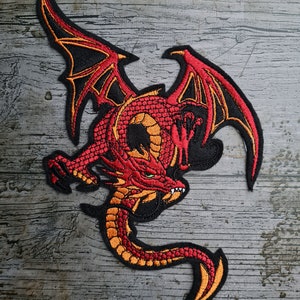 Peut inclure: Patch brodé représentant un dragon rouge et orange avec des contours noirs. Le dragon est en position de vol, les ailes déployées et la queue enroulée.