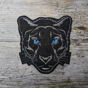 May include: Black embroidered patch of a panther's face with blue eyes. The patch is on a wood background.