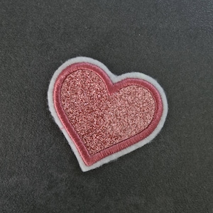 Pode incluir: Um remendo em forma de coração rosa com glitter e uma borda branca. O remendo é feito de tecido e tem uma textura felpuda.