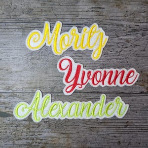 May include: Three embroidered names on a wood background. The names are Mority in yellow, Yvonne in red, and Alexander in green.