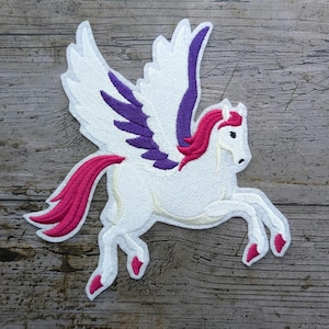 Peut inclure: Écusson brodé d'un Pégase blanc avec des accents violets et roses. Le cheval ailé a une crinière et une queue roses, et est représenté à mi-pas. L'écusson est sur un fond en bois.