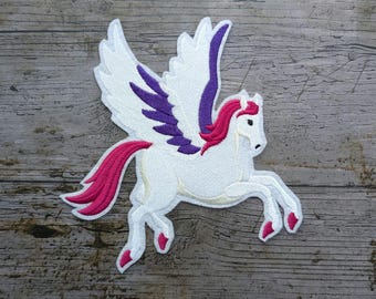 Puede incluir: Parche bordado de un Pegaso blanco con detalles morados y rosas. El caballo alado tiene una melena y cola rosas, y se muestra a medio paso. El parche está sobre un fondo de madera.