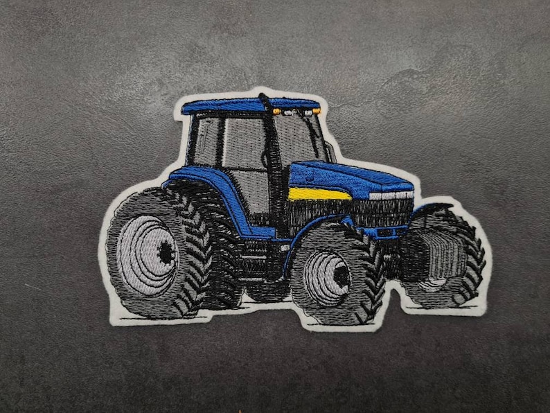 Op de afbeelding: Gestikt patch van een blauwe en gele tractor. De tractor heeft grote banden en een gedetailleerd ontwerp.