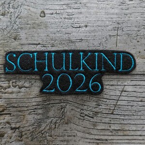 以下が含まれることがあります： 黒色の刺繍パッチ。 ターコイズ色の文字で「SCHULKIND 2026」と書かれています。 パッチは長方形で、底が湾曲しており、風化した木材の背景に配置されています。
