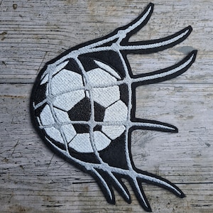 以下が含まれることがあります： サッカーボールがゴールネットに入る、黒と白の刺繍パッチ。ネットは白い糸でできており、ボールは白地に黒いステッチが施されています。
