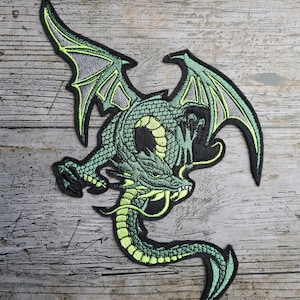 May include: Green embroidered dragon patch with black and grey accents. The dragon is in a flying pose with its wings spread out and its tail curled around its body.