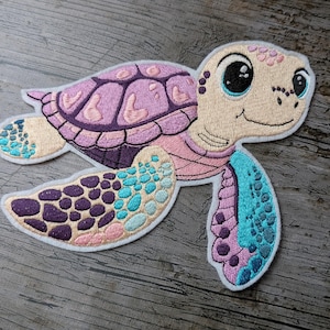May include: Embroidered patch of a smiling sea turtle in shades of pink, purple, and teal. The turtle has a light tan face and a shell with a detailed pattern. The patch is outlined in white.