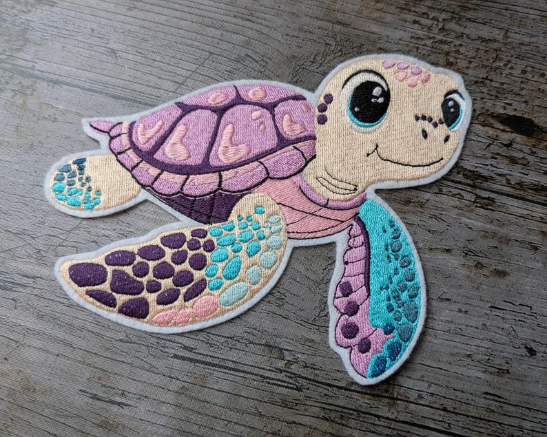 May include: Embroidered patch of a smiling sea turtle in shades of pink, purple, and teal. The turtle has a light tan face and a shell with a detailed pattern. The patch is outlined in white.