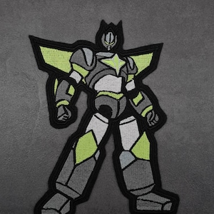 Puede incluir: Parche bordado en negro y verde de un robot con una estrella en el pecho. El robot tiene un diseño amarillo y verde en sus alas.