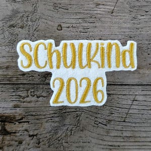 May include: Embroidered patch with the words "Schulkind 2026" in gold thread on a white background. The patch is in a stylized font and is set against a wooden background.