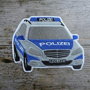 Puede incluir: Un parche bordado que representa un coche de policía alemán en azul y blanco. El coche tiene la palabra "Polizei" escrita en la parte delantera y superior del coche.