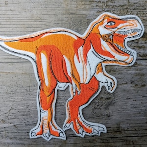 May include: Orange and white embroidered dinosaur patch. The dinosaur is a Tyrannosaurus Rex with its mouth open and teeth showing. The patch is on a white background.