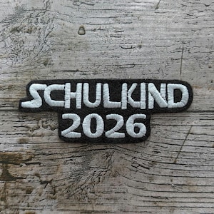 以下が含まれることがあります： 黒い背景に白い糸で「SCHULKIND 2026」と刺繍されたパッチ。 パッチは角が丸い長方形で、風化した木材の表面に配置されています。