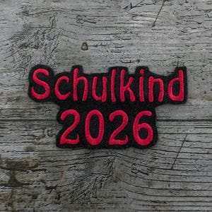 May include: A black patch with the words "Schulkind 2026" in red lettering. The patch is on a weathered, grey wooden surface. The text is in a stylized font.