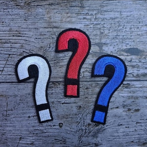 Könnte beinhalten: Drei Fragezeichen-Patches in Weiß, Rot und Blau mit schwarzen Umrissen. Die Patches sind gestickt und haben einen schwarzen Rand. Die Patches sind auf einer verwitterten, grauen Holzoberfläche angeordnet.