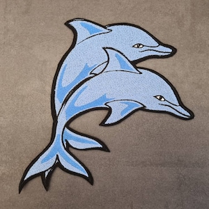Op de afbeelding: Twee blauwe geborduurde dolfijn patches, een bovenop de andere, met zwarte contouren.
