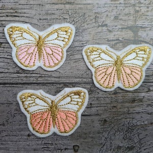 Op de afbeelding: Drie roze en witte geborduurde vlinderpatches met gouden accenten.