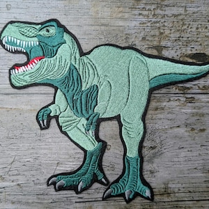Peut inclure: Un patch thermocollant brodé représentant un dinosaure Tyrannosaurus Rex vert avec une gueule ouverte montrant des dents.