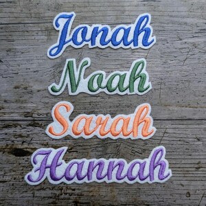 Könnte beinhalten: Gestickte Namensaufnäher in Blau, Grün, Orange und Lila. Die Namen Jonah, Noah, Sarah und Hannah sind in einer Schreibschrift mit weißem Rand auf einem verwitterten Holzgrund dargestellt. Ideal zum Personalisieren von Artikeln.