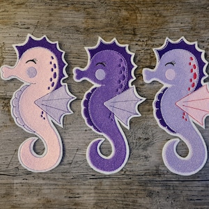 Puede incluir: Tres parches bordados de caballitos de mar en tonos morados y rosas. Cada caballito de mar tiene un contorno blanco, una aleta detallada y una cara sonriente. Los caballitos de mar se muestran sobre una superficie de madera.