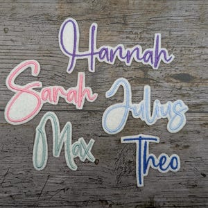 Könnte beinhalten: Eine Sammlung von Namensaufnähern aus Stoff in verschiedenen Farben. Die Namen sind "Hannah" in Lila, "Sarah" in Rosa, "Julius" in Hellblau, "Max" in Türkis und "Theo" in Blau. Jeder Name hat einen weißen Hintergrund und eine farbige Umrandung.