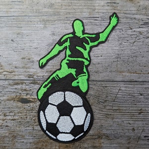 May include: Green and black embroidered patch of a soccer player kicking a soccer ball.
