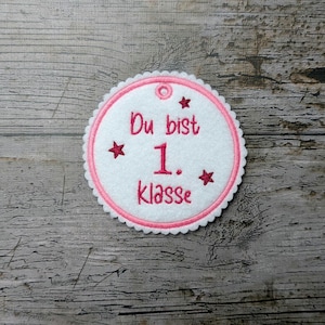 Op de afbeelding: Een witte vilten patch met een roze geschulpte rand en een roze geborduurd ontwerp. De tekst "Du bist 1. Klasse" is roze, met steraccenten. Er zit een klein gaatje aan de bovenkant om op te hangen.