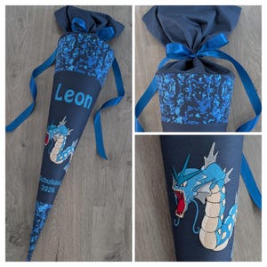 Könnte beinhalten: Eine dunkelblaue Schultüte mit blauem Tarnmuster und blauem Band. Die Tüte trägt den Namen "Leon" in Hellblau und einen gestickten blau-weißen Drachen. Der Text "Schulkind 2026" ist ebenfalls sichtbar.