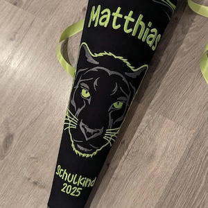 May include: A black and green school cone with a black panther face and the name "Matthias" embroidered in green. The cone also has the text "Schulkind 2025" embroidered in green.