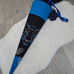 Könnte beinhalten: Ein blauer und schwarzer Schultüte mit einem schwarzen Panthergesicht und silbernen Krallen. Die Tüte hat ein blaues Band und ein Pfotenanhänger mit dem Jahr 2022.