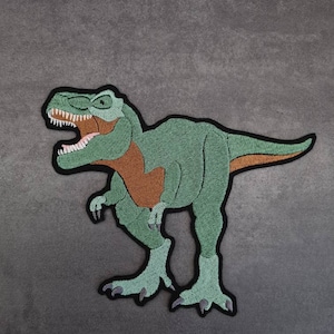 May include: Green embroidered dinosaur patch with black stitching and brown accents. The dinosaur is standing on its hind legs with its mouth open and teeth showing.