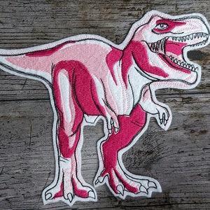 May include: Pink embroidered iron-on patch of a Tyrannosaurus Rex dinosaur with white outline.