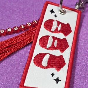 May include: Embroidered keychain with a red and white design. The rectangular tag features three red shapes with white accents, a red border, and two black star shapes. A red tassel and beaded strand with the letters "LET PRO" are attached.