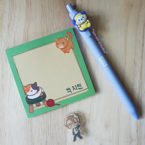 May include: A light blue pen with a yellow cartoon character, a green sticky note pad with cat illustrations, and a small cartoon pin of a man. The pen has the text "* pen not included *".