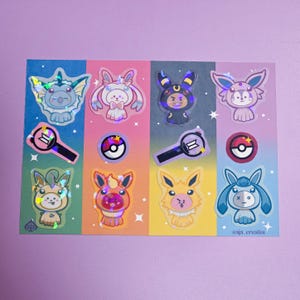 May include: A rectangular sticker sheet featuring holographic illustrations of cartoon characters in animal costumes, small circular icons, and light sticks. The background is divided into four vertical colour panels. The text "aja_creates" is printed in the bottom right corner.