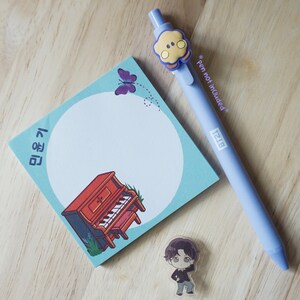May include: A light blue pen with a yellow and blue character, a light blue sticky note with a piano illustration, a purple butterfly, and a small character figurine on a wooden surface.