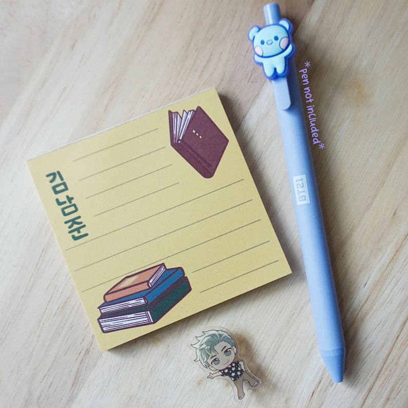 May include: A light blue pen with a cartoon bear, a yellow notepad with book illustrations and Korean text, and a small cartoon figure on a wooden surface. The pen has "BT21" printed on it.