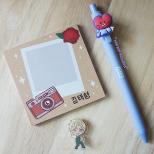 May include: A light blue pen with a BT21 character topper, a beige polaroid-style notepad with a camera illustration, a red rose, and a small acrylic figure of a cartoon character. The notepad has Korean text.
