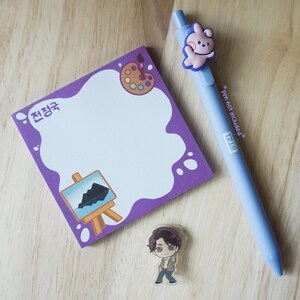 May include: A purple notepad with a white, cloud-shaped space for writing, featuring a paint palette and easel illustration. A light blue pen with a cartoon character topper and a small acrylic figure are also present.