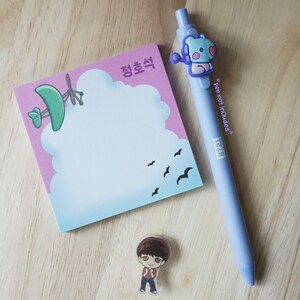 May include: A light blue pen with a cartoon character, a pink notepad with a cloud design and Korean text, and a small cartoon figure on a wooden surface.