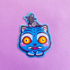 May include: A blue cat sticker with large orange eyes, pink cheeks, and a small hat. The cat has a wide grin and small paws. The sticker has a light blue outline and is on a purple background.