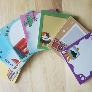 May include: A selection of square sticky notes featuring illustrations and Korean text. Designs include a fishing rod, piano, cat, camera, and paint palette. The notes are arranged on a light wood surface.